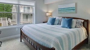 A bedroom with a large window, a bed with blue and white bedding, a nightstand, and a painting on the wall. - Make your summer memories here! (Wells)