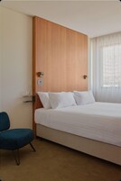 Una camera d'albergo con un letto, una poltrona blu, una testiera in legno, un comodino e una finestra con tende.