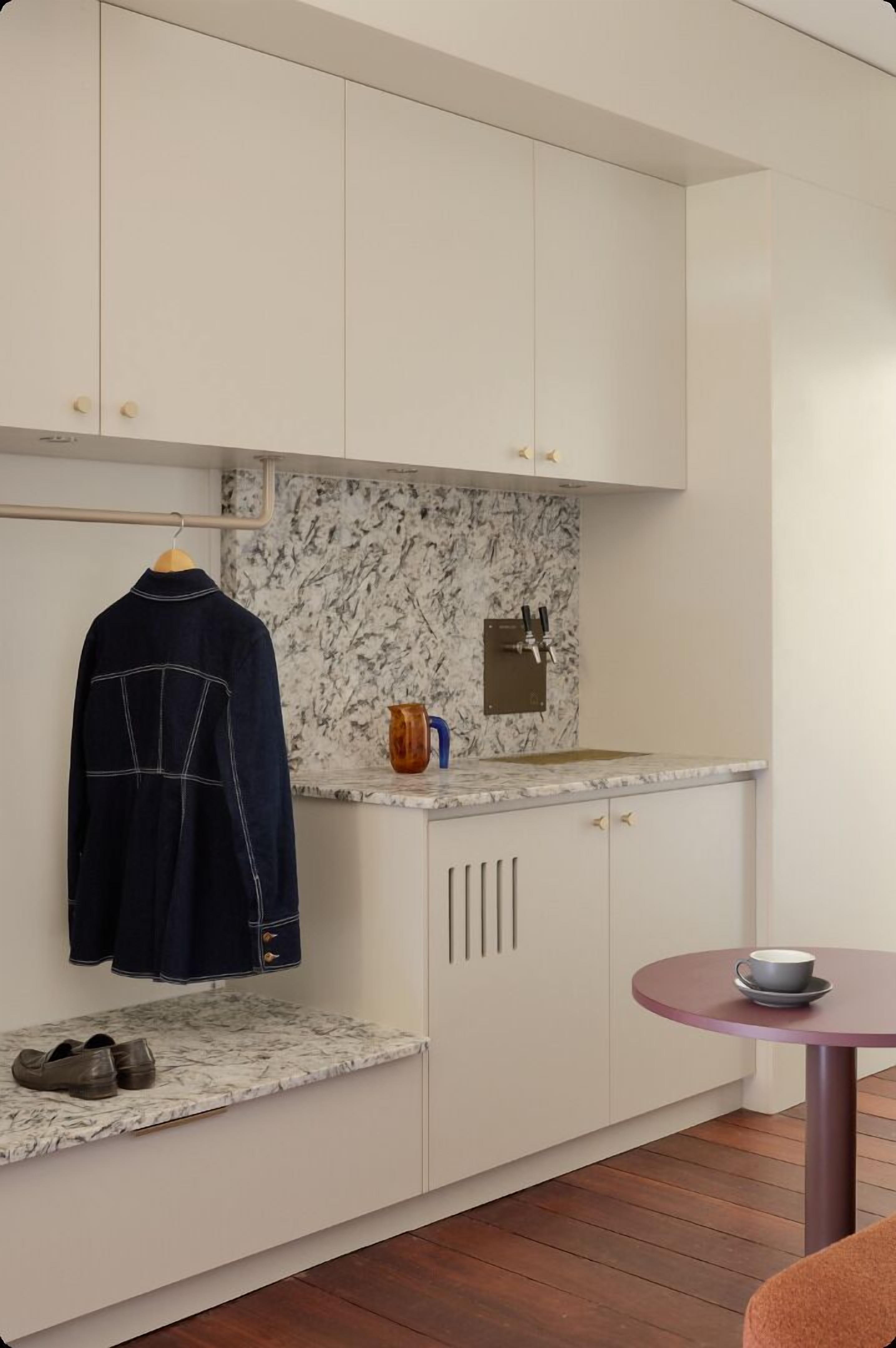 Une cuisine compacte avec des meubles blancs, un pare-feu en marbre, un évier avec un robinet, une petite table avec un saladier et un porte-manteau suspendu.
