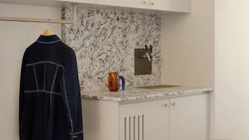 Cocina compacta con armarios blancos, salpicadero de mármol, un lavabo con grifo, una mesita con un bol y un perchero.