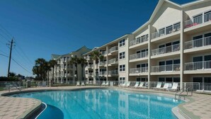 Una piscina con agua cristalina azul, rodeada por un edificio de varios pisos con balcones y un camino empedrado.