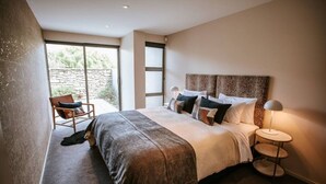 A bedroom with a large bed, a chair, a lamp, and a view of the outdoors. - Lake Edge | Central lakefront apartment | 3 bed (Wanaka)
