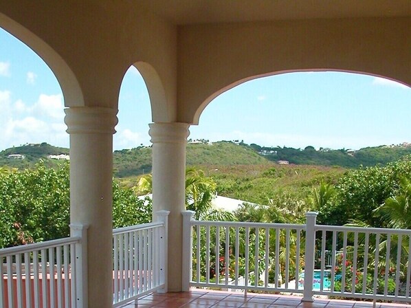 Ein überdachter Balkon mit weißen Geländern, der einen Blick auf eine üppige Landschaft mit Hügeln und viel Grün bietet.