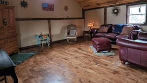 A cozy loft-style room with wooden floors, a leather sofa, a wooden cabinet, and a small desk with a chair. - The Barn House Lodge (Spillville)
