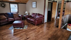 A cozy living room with a wooden ceiling, a brown leather sofa, a matching armchair, and a wooden floor. - The Barn House Lodge (Spillville)