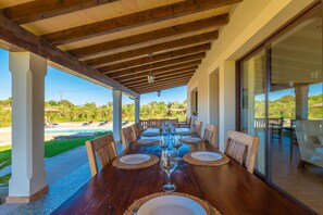 A covered dining area with a wooden table set for six, overlooking a pool and greenery.