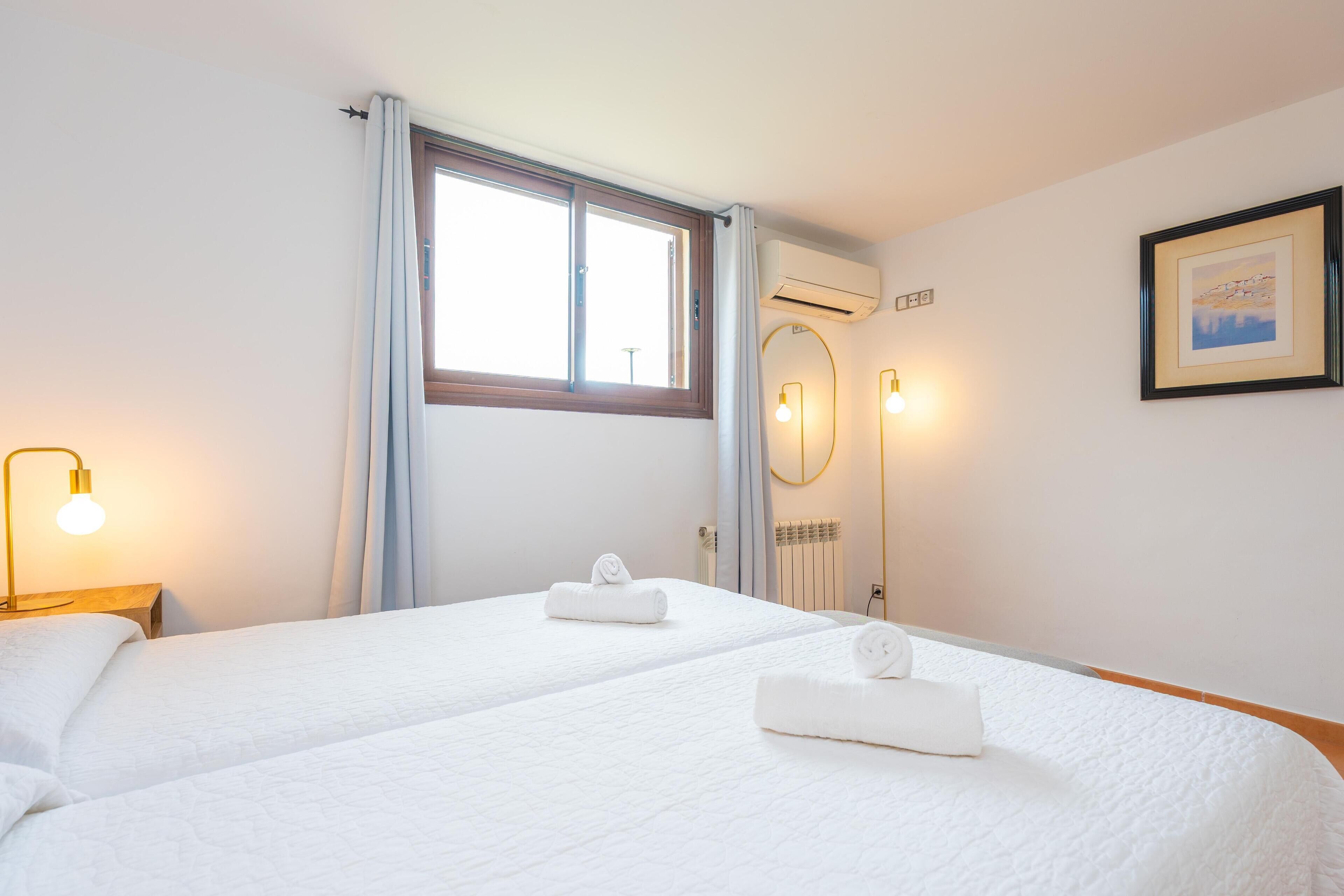 A hotel room with two beds, a window with curtains, an air conditioner, a framed picture on the wall, and a bedside lamp.