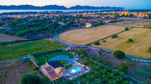 An aerial view of a residential area with a swimming pool, a curved road, and surrounding fields.