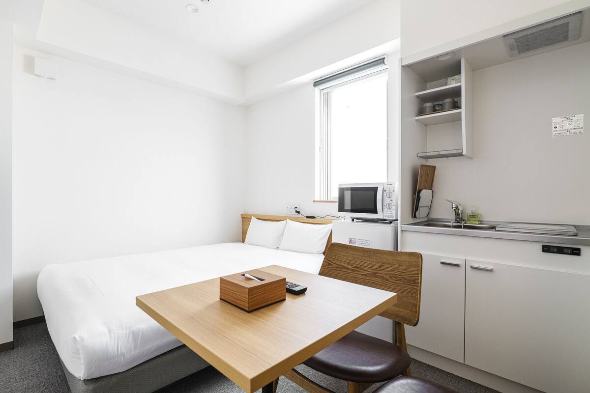 A hotel room with a bed, a wooden table with a box and remote, a chair, a kitchenette with a microwave, and a window.