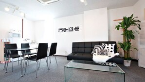 Un salon moderne avec une table basse en verre, un canapé noir et un coin repas comprenant des chaises, avec une cuisine en arrière-plan.