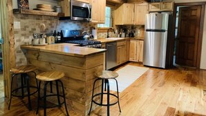 Una cocina con gabinetes de madera, un refrigerador de acero inoxidable, un microondas y una estufa. Hay dos taburetes y un fregadero.