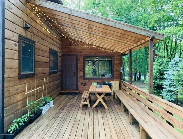 Relax on the covered porch with a wooden table and benches, surrounded by lush greenery and string lights.