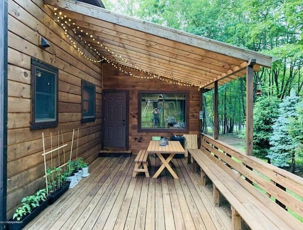 Relax on the covered porch with a wooden table and benches, surrounded by lush greenery and string lights.