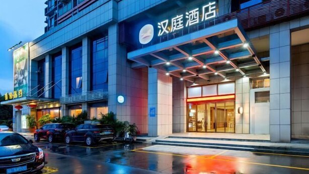 A hotel entrance with a modern facade, a visible sign indicating the hotel's name, and a parking area with cars. - Hanting Hotel (Guangzhou Feixiang Park Metro Station) (Guangzhou)