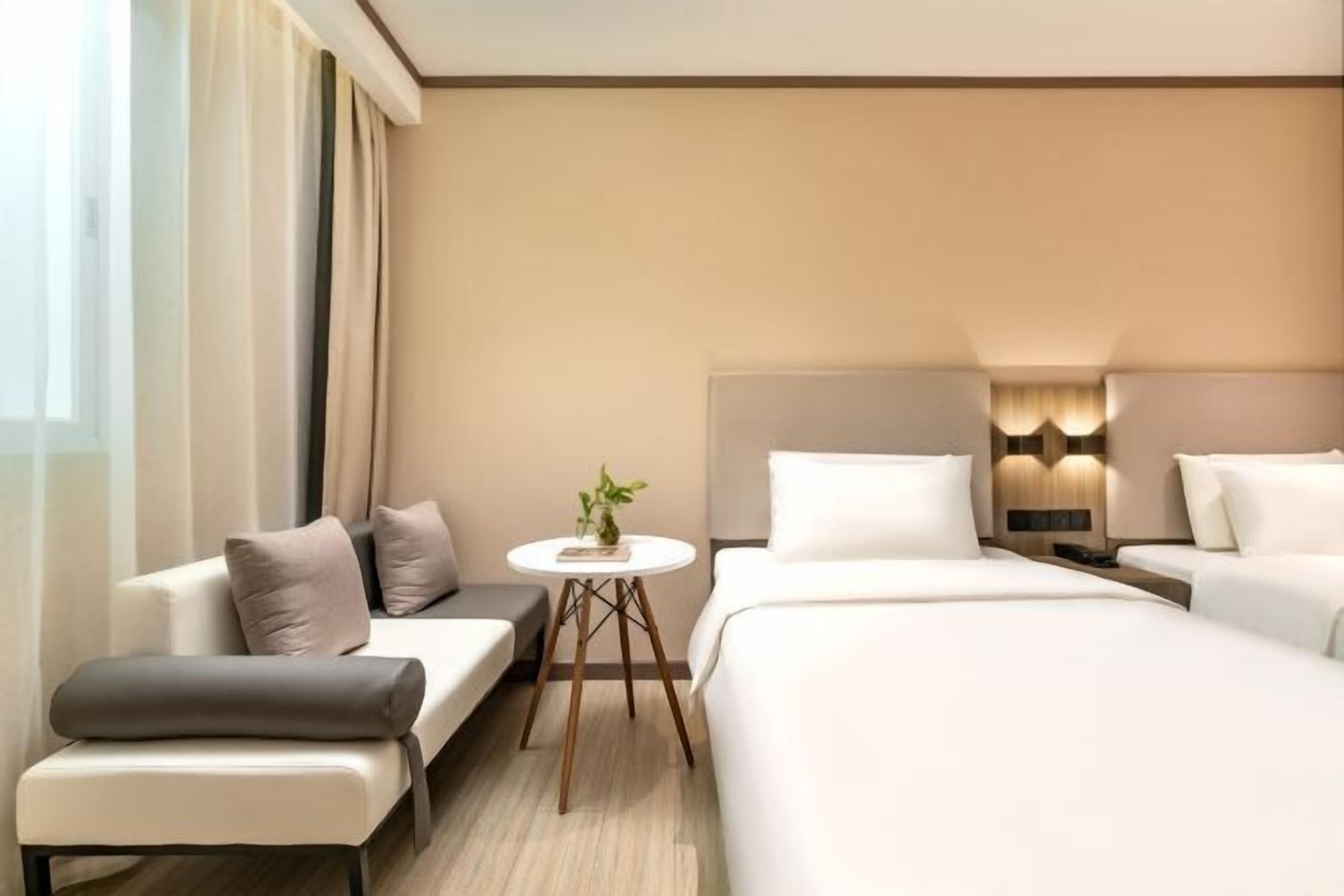 Een moderne hotelkamer met een bank, twee enkele bedden, een klein rond tafeltje met een plant en een wandlamp.