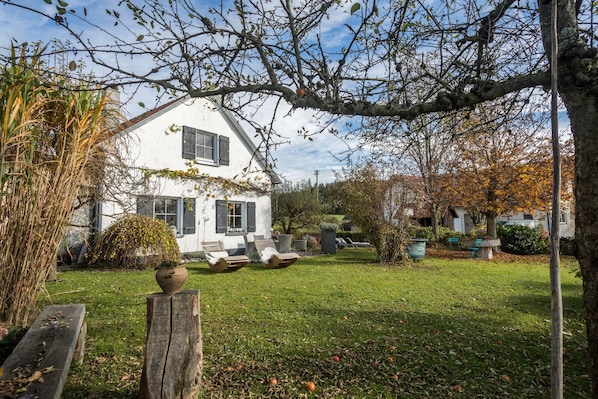 A white house with a sloped roof, a garden with grass, a wooden bench, and a tree with bare branches.