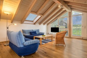 A modern living room with a blue sofa, a wicker chair, a coffee table, and a TV. - Beautiful Top Floor Holiday Apartment "Schleinseeblick" with Lake View (Kressbronn)