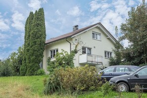 Une maison à deux étages avec un balcon, entourée de verdure et avec une voiture garée devant.
