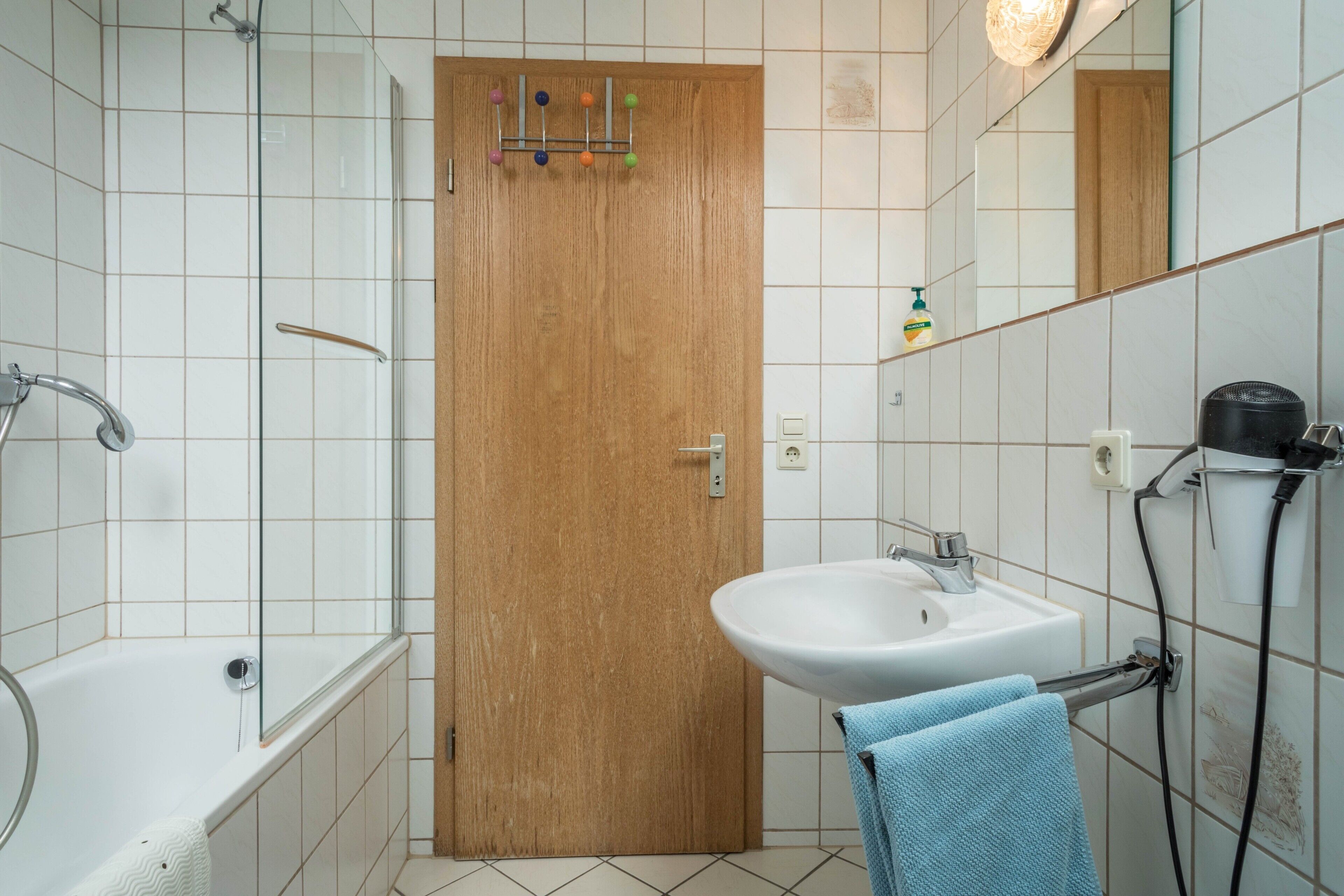 Un bany amb una porta de fusta, un lavabo blanc, una banyera i un penjador de tovalloles.