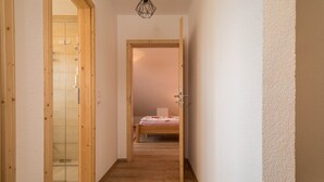 Ein enger Korridor mit Holzboden, einer Duschkabine aus Glas und einem Bett mit rosa Bettdecke im Hintergrund.