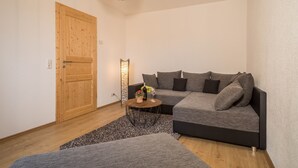 Ein modernes Wohnzimmer mit Holztür, grauem Sofa, kleinem runden Tisch mit Flasche und Glas sowie einem gemusterten Teppich.