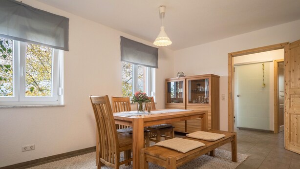 Ein Essbereich mit Holztisch, Stühlen, einer Bank, einer Blumenvase und einer Pendelleuchte.
