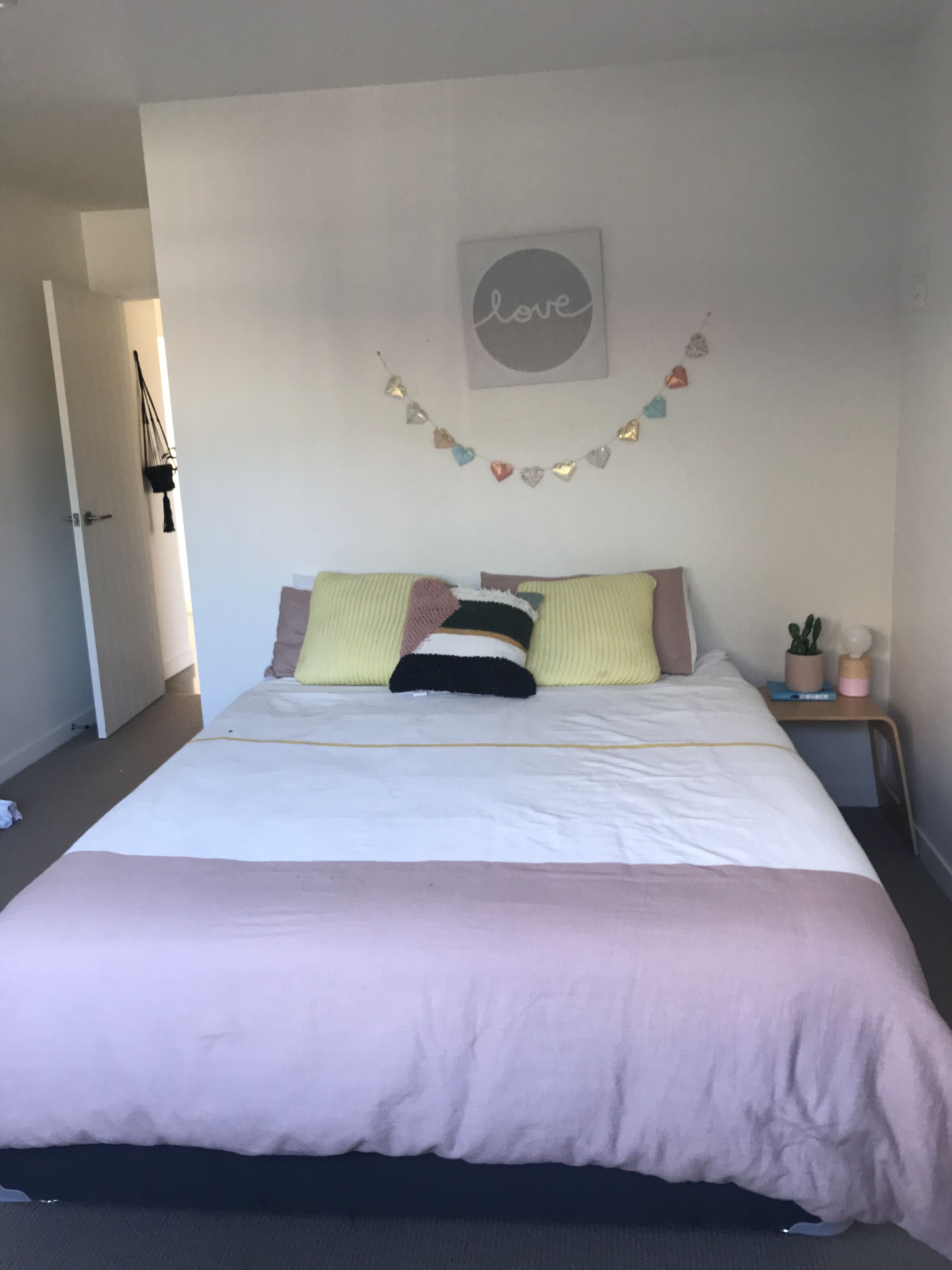 Ein ordentlich gemachtes Bett mit einer rosa Decke, einer herzförmigen Wanddekoration und einem kleinen Nachttisch mit einer Pflanze.