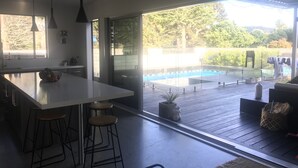 A modern kitchen with a large island, bar stools, and a view of an outdoor swimming pool. - Luxury Home with pool/spa; close to beach/ 4 bedroom/ 2 bath. On golf course. (Whangamata)