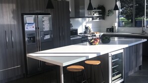 A modern kitchen with a large island, bar stools, stainless steel appliances, and a view of the outdoors. - Luxury Home with pool/spa; close to beach/ 4 bedroom/ 2 bath. On golf course. (Whangamata)