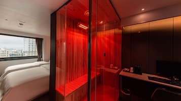 A modern hotel room with a large bed, a desk with a computer, and a view of the city through a large window.