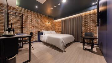 A modern hotel room with a brick wall, a bed, a desk with a chair, a small table, and a TV mounted on the wall.