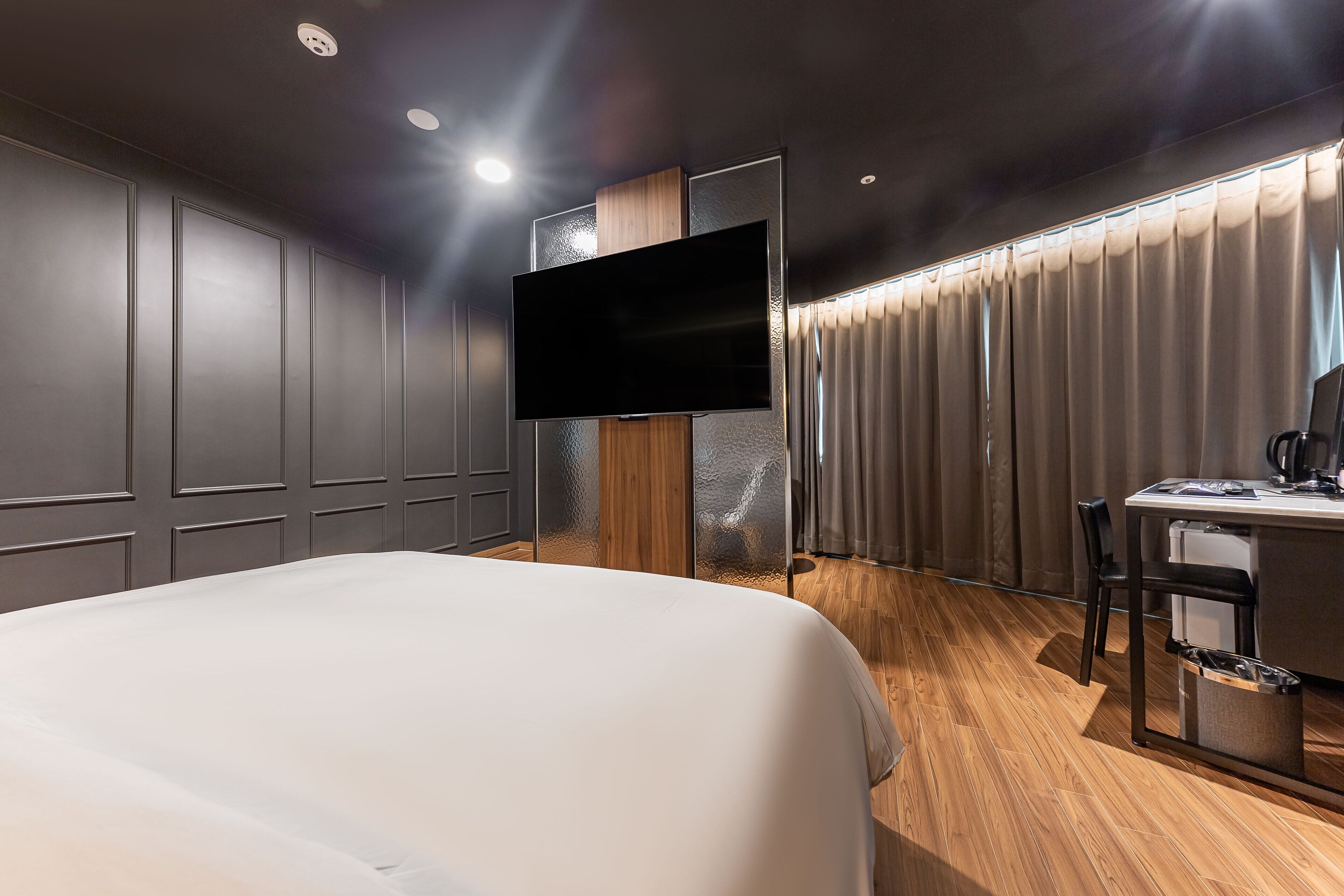 A modern hotel room with a large bed, a flat-screen TV mounted on the wall, a desk with a chair, and a window with curtains.