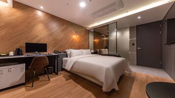 A modern hotel room with a wooden floor, a bed with white linens, a desk with a computer, and a small refrigerator.