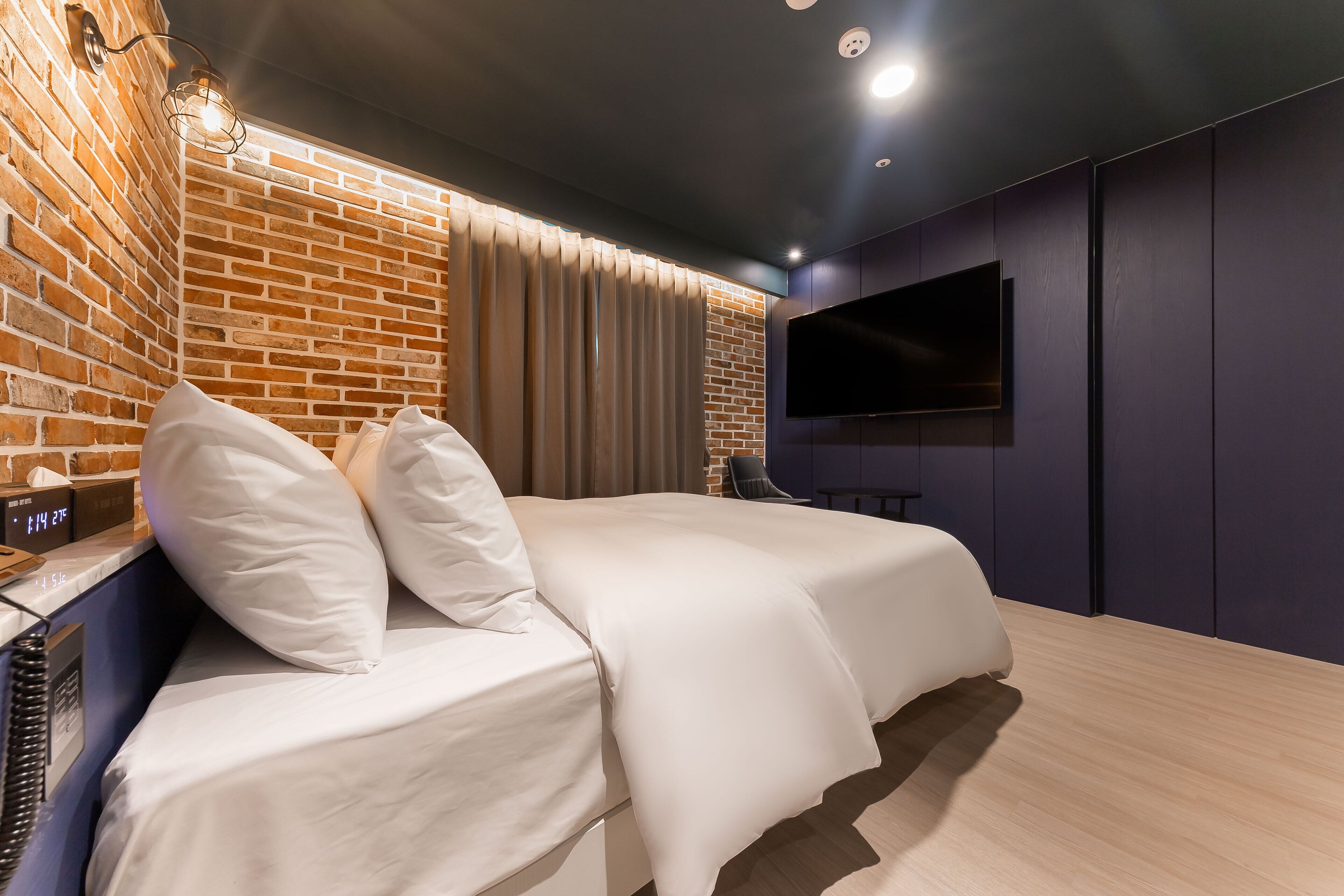 Quarto de hotel moderno com parede de tijolos decorativa, uma TV de tela plana fixada na parede e uma cama com roupa de cama branca.