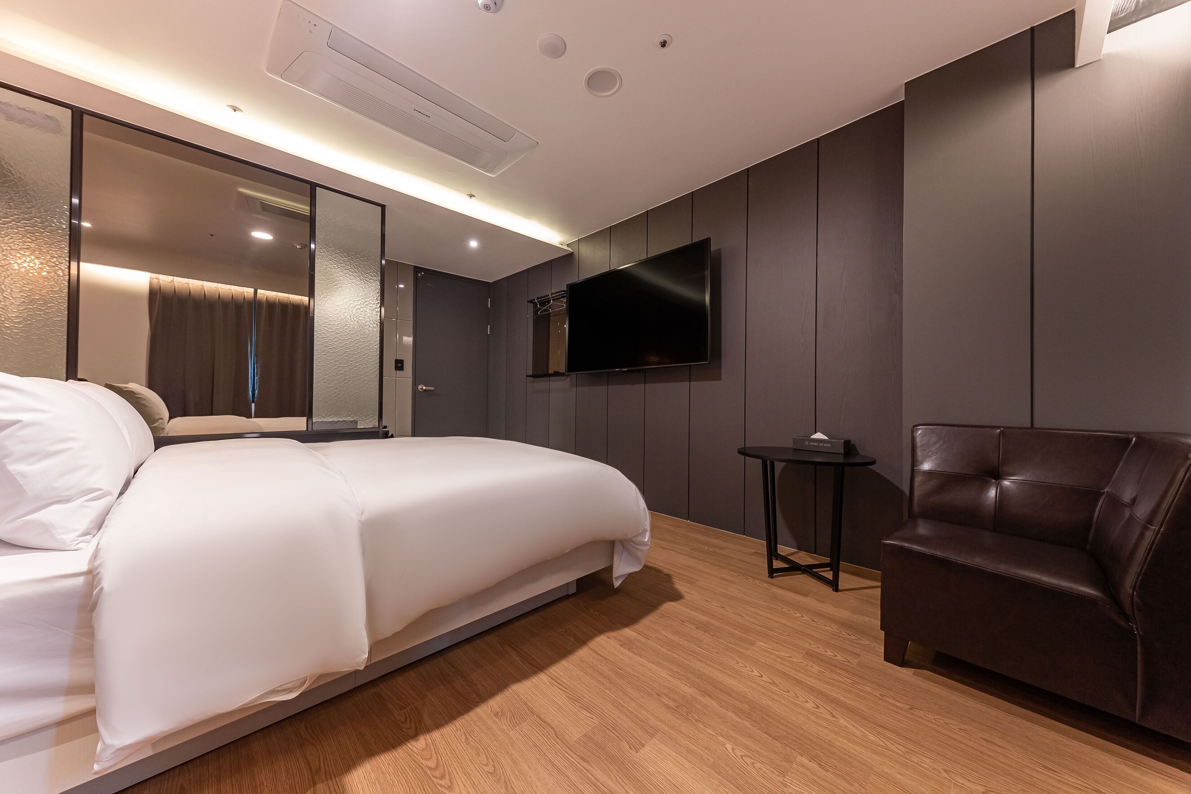 Una habitación de hotel moderna con una cama grande, un televisor de pantalla plana montado en la pared, un sillón de cuero y una pequeña mesita.