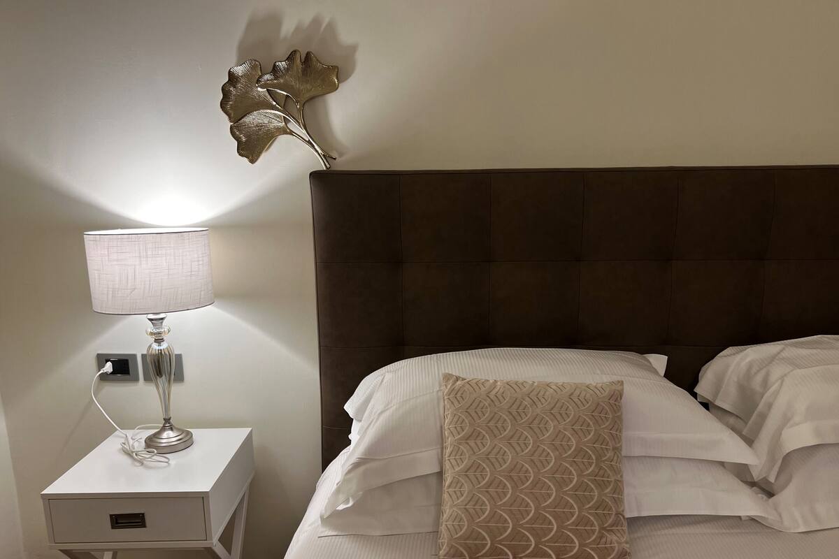 A neatly made bed with white linens and a patterned pillow, a bedside lamp on a nightstand, and a decorative wall piece.