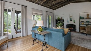 Een gezellige woonkamer met een blauwe bank, een houten tafel met een lamp, een open haard en uitzicht op groen.