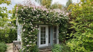 Een klein, wit huisje met een groene deur, omringd door weelderige begroeiing en roze bloemen.