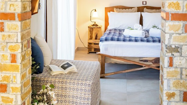 Un dormitorio con un arco de ladrillo, una cama con ropa de cama blanca, una mesita de noche con una lámpara, y un pequeño estante con objetos decorativos.