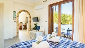 Habitación de hotel con una cama grande, televisión, balcón con mesa y sillas, y un arco de ladrillo que conduce al comedor.