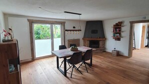 A spacious living room with a wooden floor, a dining table with chairs, a fireplace, and a bookshelf.