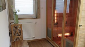 A room with a wooden floor, a window with blinds, a radiator, a wooden table, and a sliding glass door leading to a sauna area.