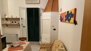 Une petite pièce, de taille modeste, comprenant un coin cuisine, un lit recouvert d’une courtepointe à motifs floraux, une table avec une nappe et une porte dotée d’un volet vert.