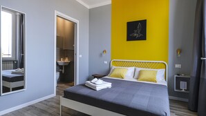 A modern bedroom with a bed, a bedside table, a mirror, and a bathroom visible through an open door. - albinariobrescia  (Brescia)