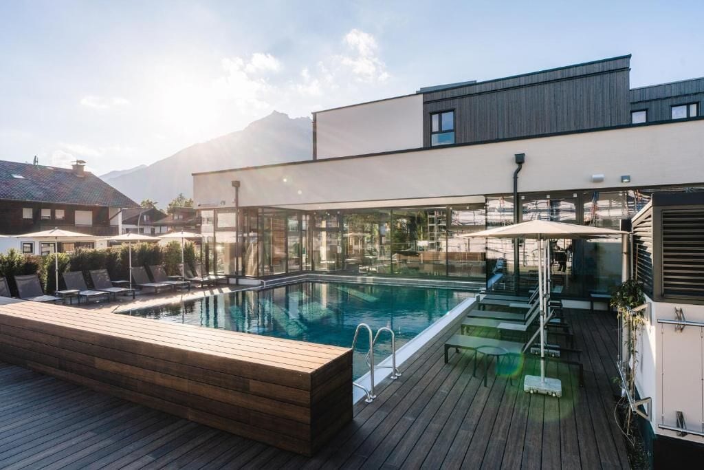 A rooftop swimming pool with a wooden deck, glass walls, and a mountain backdrop.