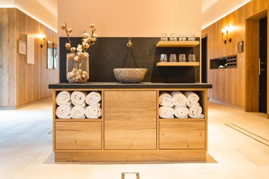 A wooden vanity with a stone basin, a vase with dried flowers, and neatly rolled towels.
