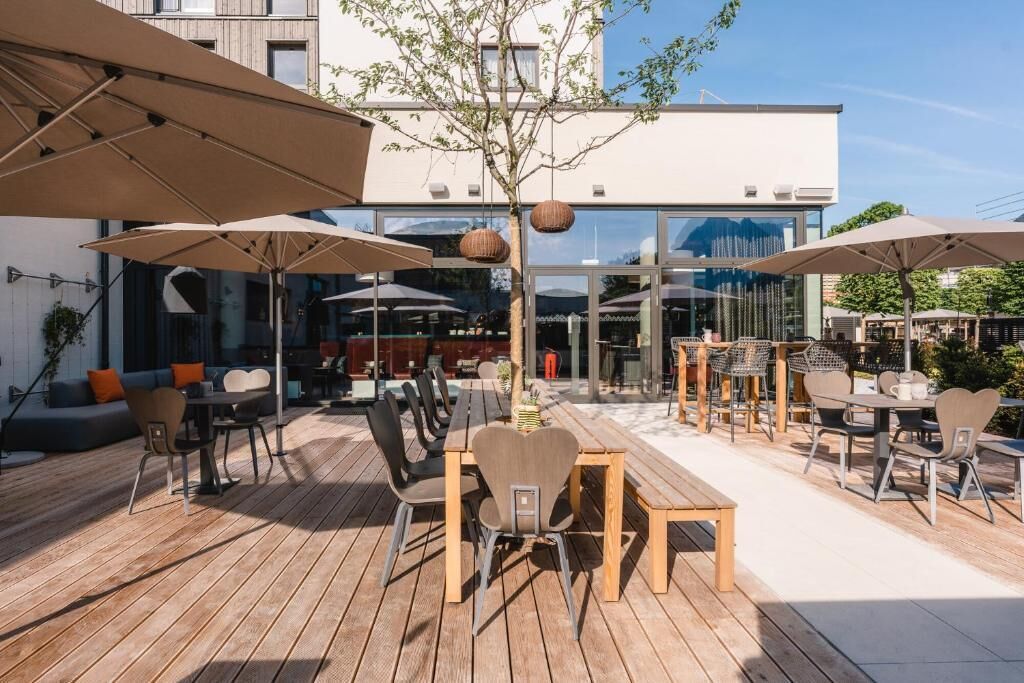 Un patio al aire libre con mesas y sillas de madera, sombrillas y vistas a un edificio moderno al fondo.