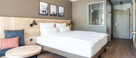 A modern hotel room with a large bed, a small round table, and a sofa. There are three framed pictures on the wall above the bed.