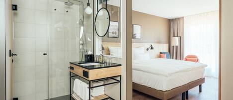 Habitación de hotel moderna con ducha envuelta en vidrio, tocador de madera con lavamanos, cama grande con ropa blanca y mesita de noche con lámpara.