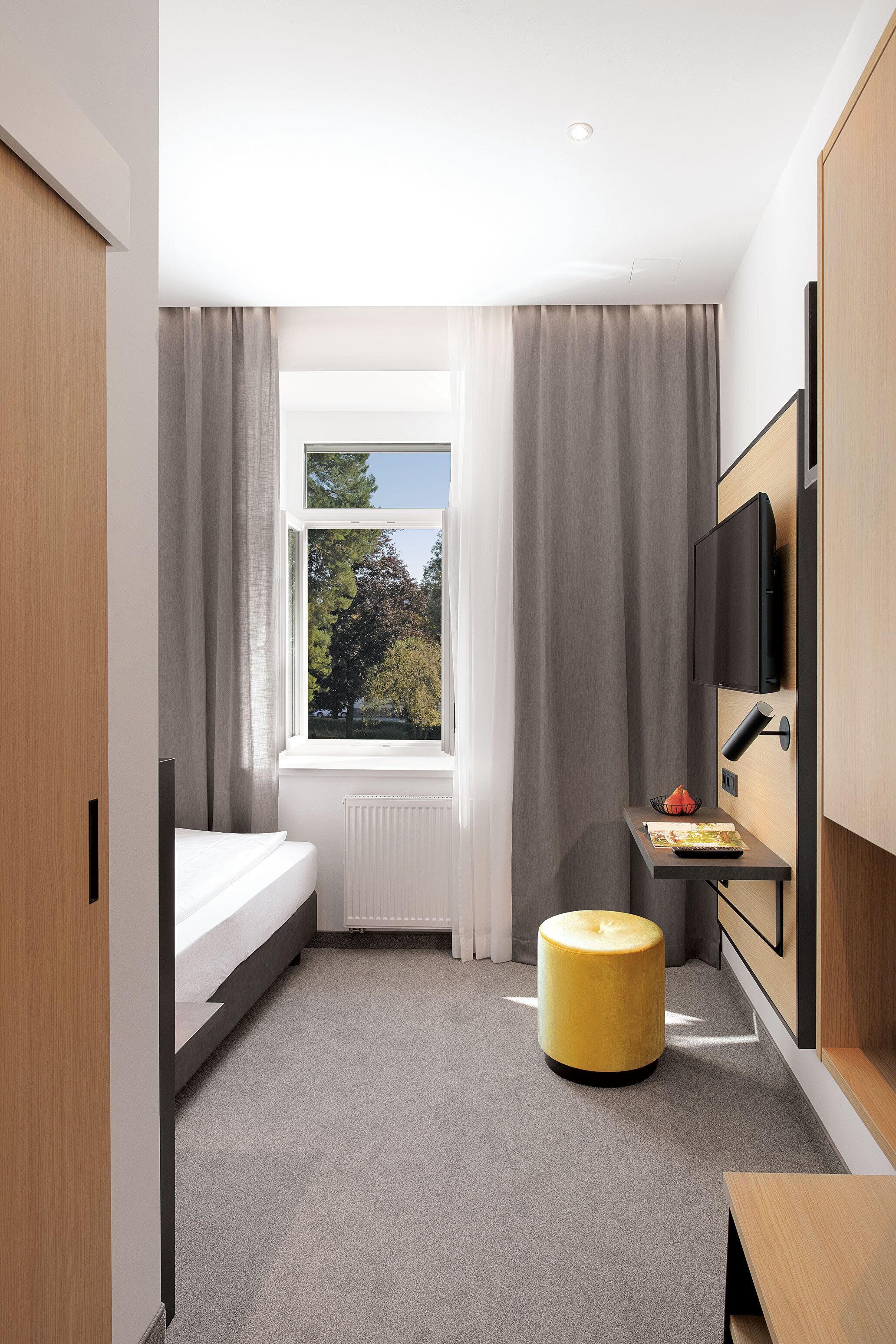 Habitación de hotel compacta con cama, escritorio, silla, televisor y ventana con cortinas.